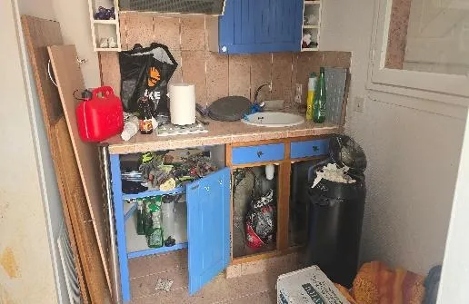 Meuble évier bleu aux portes ouvertes dans une petite cuisine encombrée, avec un bidon rouge, des bouteilles et une poubelle noire