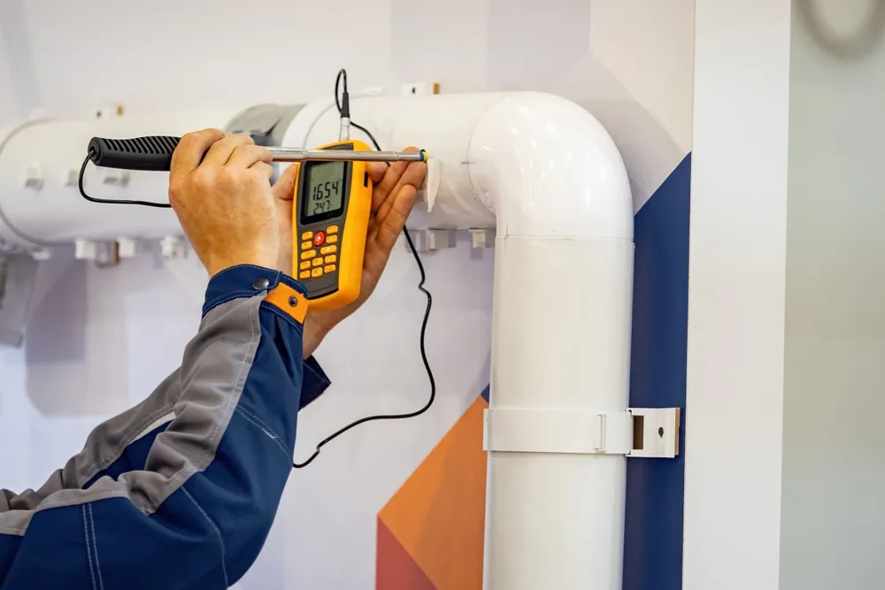 Technicien en tenue bleue et grise mesurant un tuyau blanc avec un appareil portable jaune muni d'une sonde